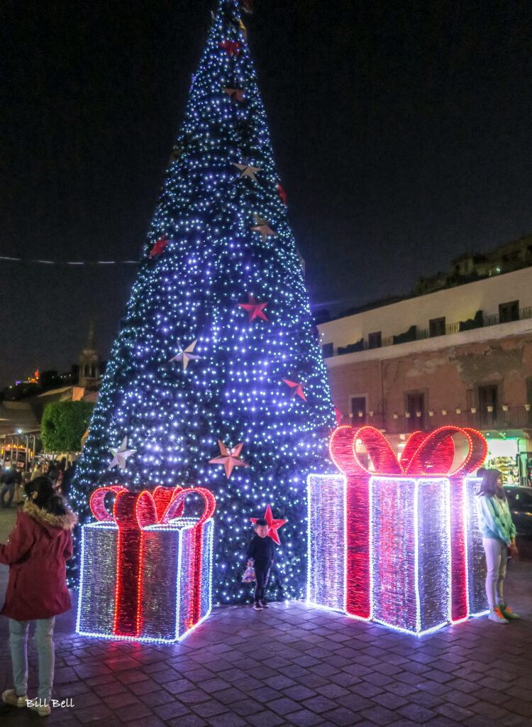 A towering Christmas tree adorned with lights and oversized gift decorations brightens a public square, merging global holiday customs with Mexico’s festive flair.