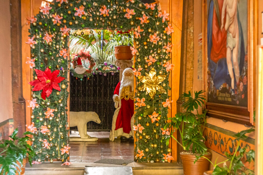 A whimsical scene featuring Santa Claus peeking out from a poinsettia-adorned doorway, blending global Christmas symbols with Mexico’s vibrant holiday decorations.